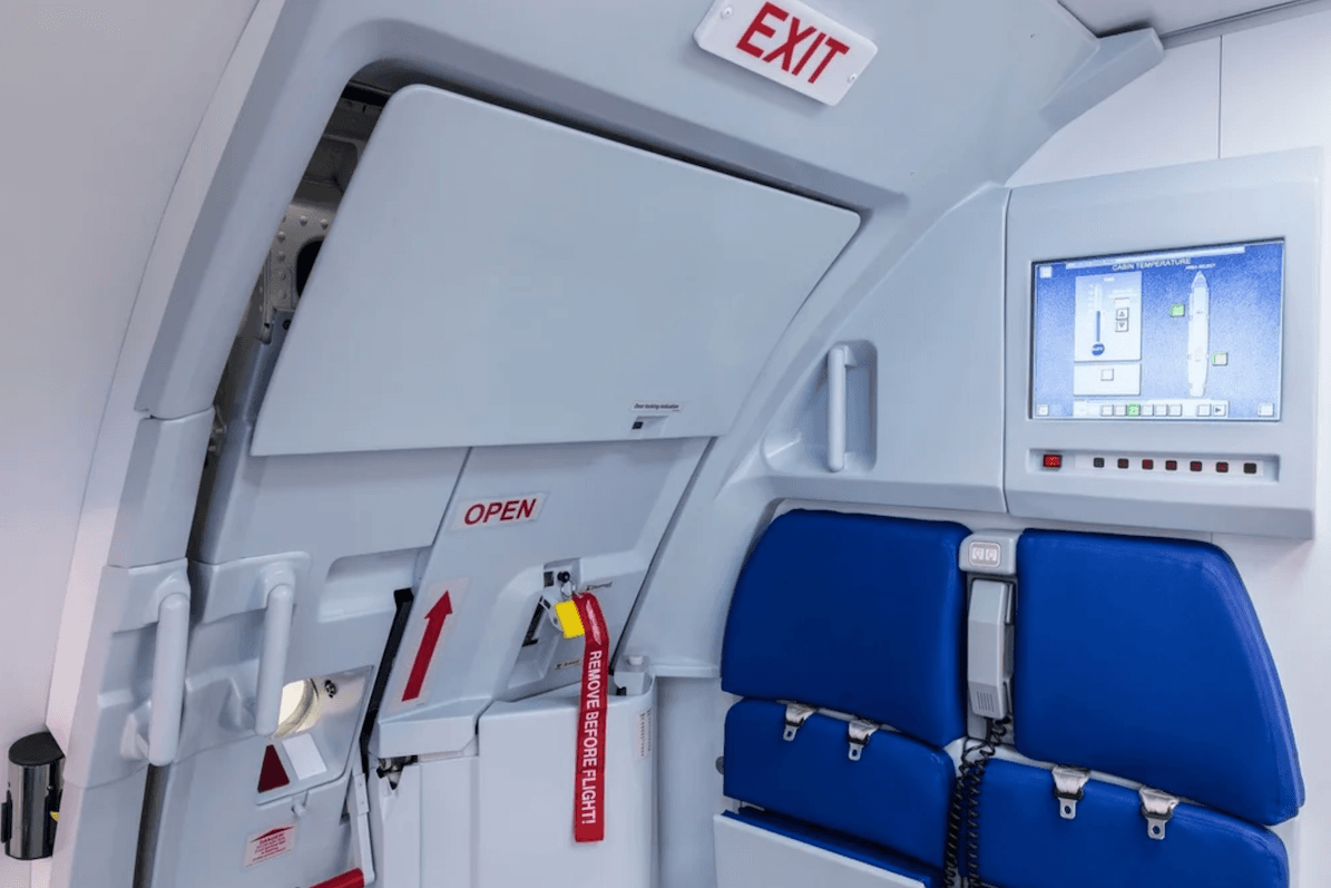 The interior of an A320 emergency exit door in a training environment. A bold red EXIT sign is mounted above the door. The word OPEN appears in red on the door mechanism, accompanied by red arrows pointing upward. A yellow remove-before-flight tag is attached to the handle.