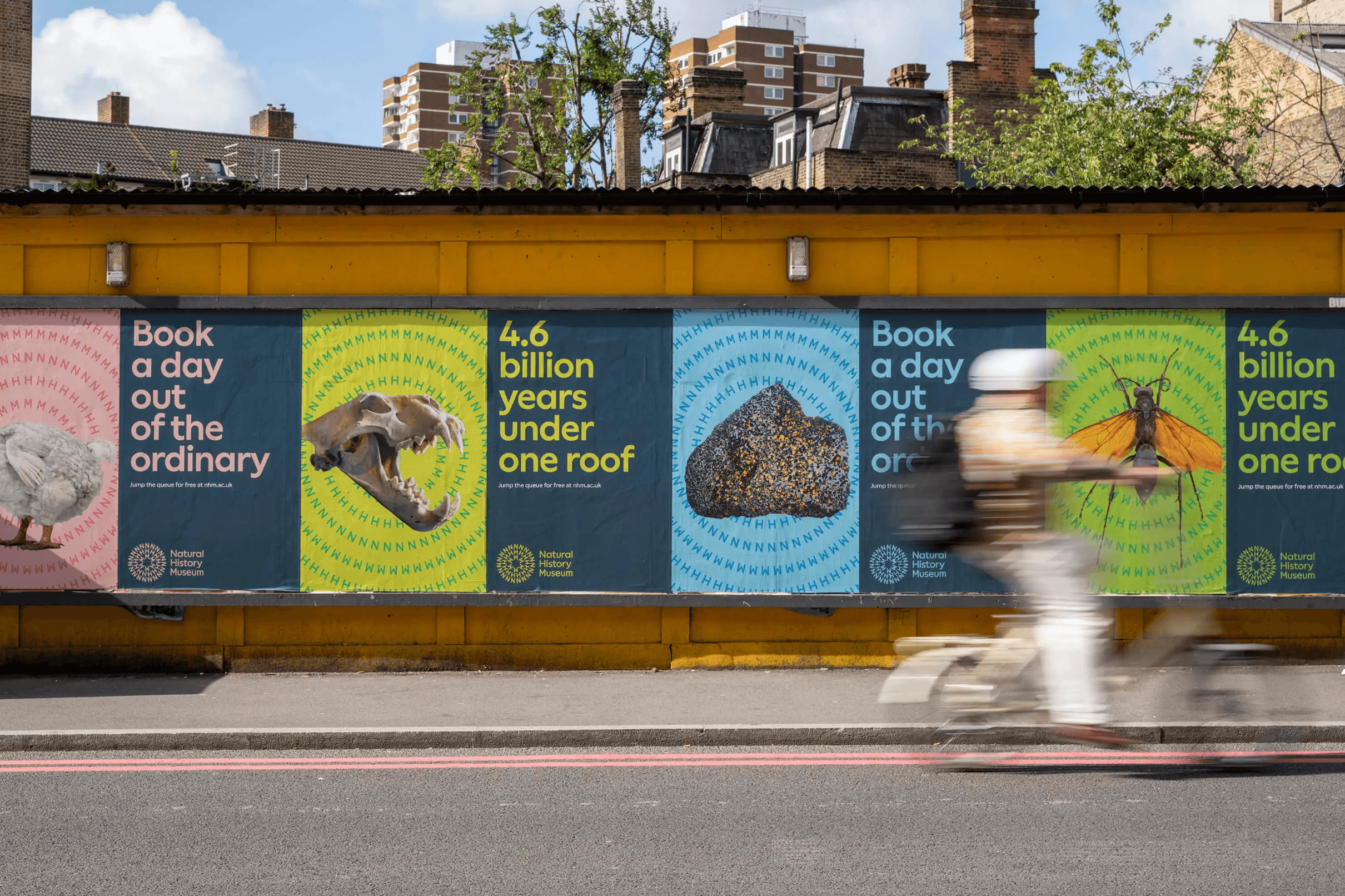 A row of NHM billboard advertisements on a London street, featuring bold colourful designs with slogans like 'Book a day out of the ordinary' and '4.6 billion years under one roof'. A cyclist blurs past in the foreground.